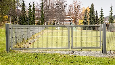 Palloilukentän ympärillä vaaleanharmaa elementtiaitaus, jossa epäsymmetrinen, saranoitu huoltoportti kääntösalpalukituksella ja maasalvoilla varustettuna.
