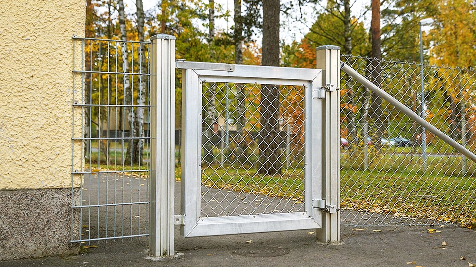 Matala, alumiininen ja panssariverkkotäytteinen yksilehtinen käyntiportti koulurakennuksen nurkalla.
